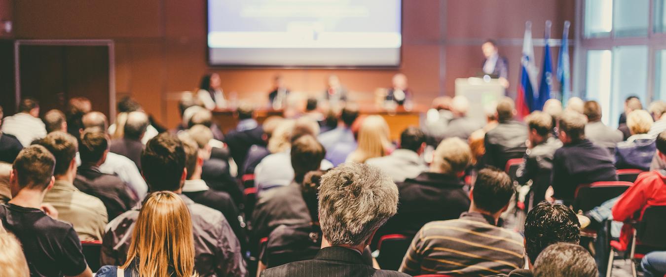 Audience in event room listens to speakers on stage give presentation