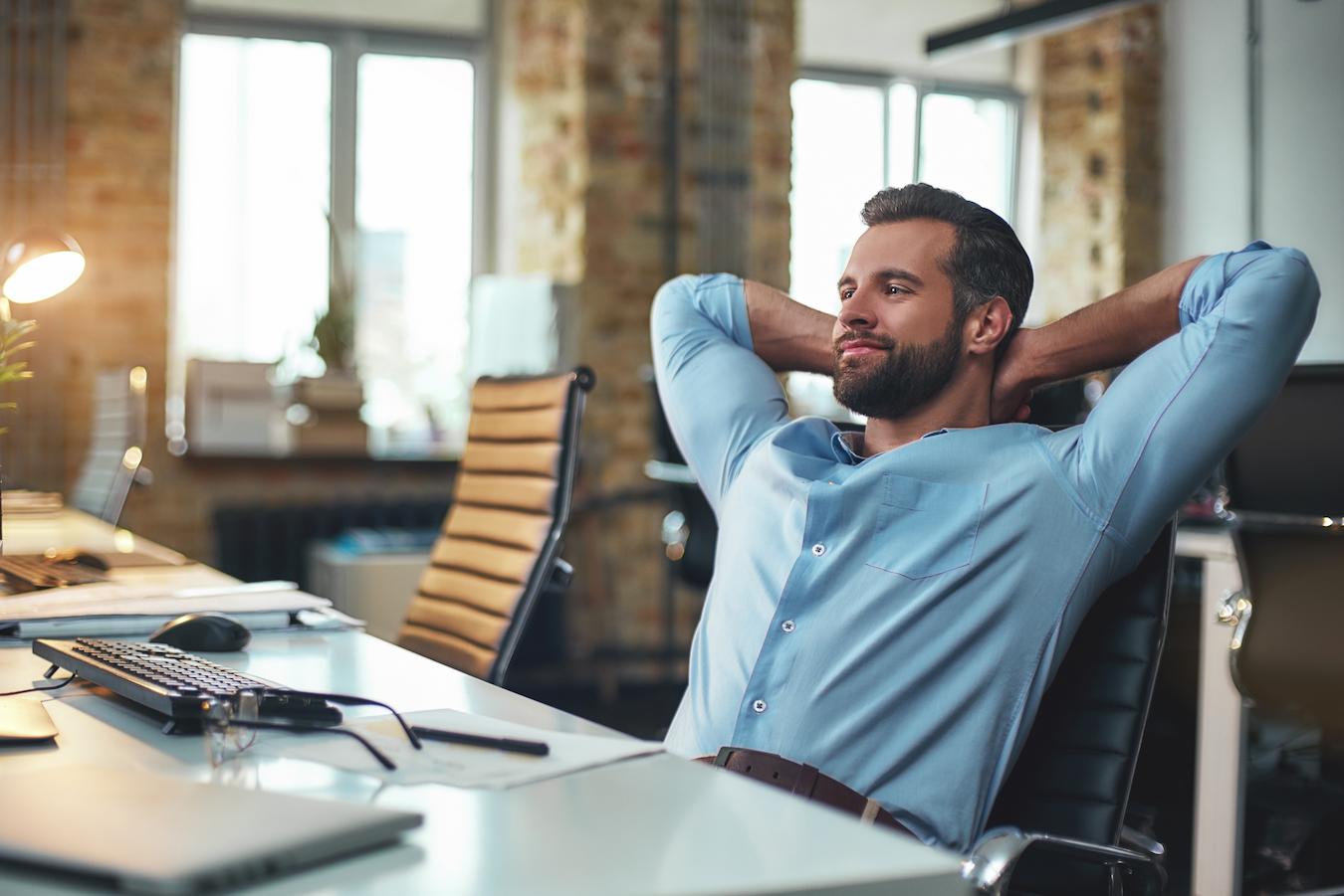 Bearded person in a blue button down shirt leans back in an office chair and puts hands behind their head while smiling in satisfaction at a computer screen