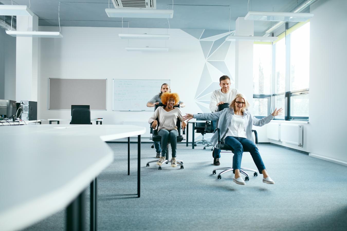 Colleagues laugh and race through office in rolling chairs