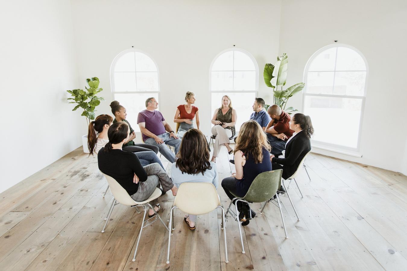 Eleven people sit in a circle of chairs in a bright room
