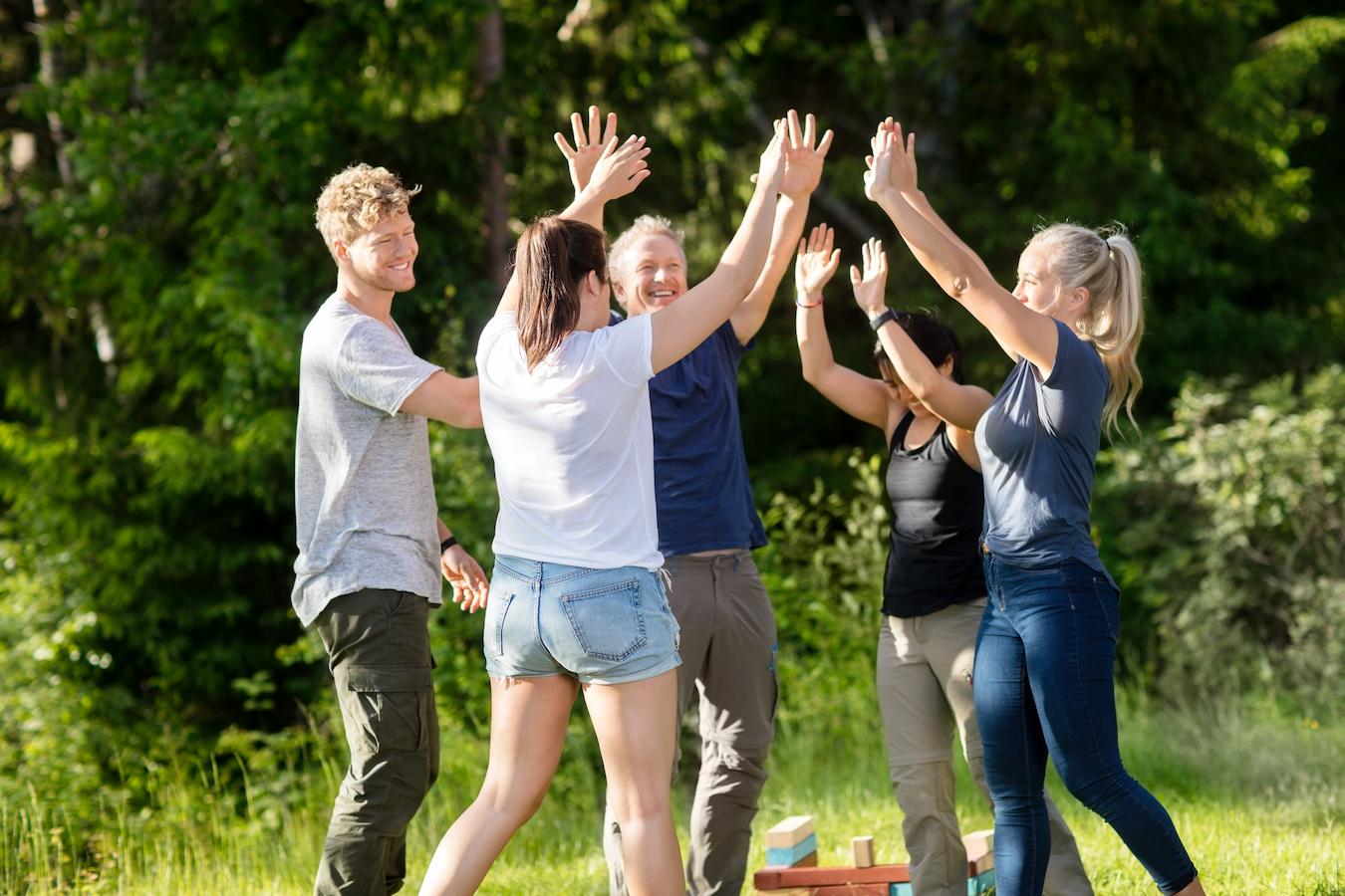 Five colleagues high five and celebrate outside in the sunshine after a successful team building exercise
