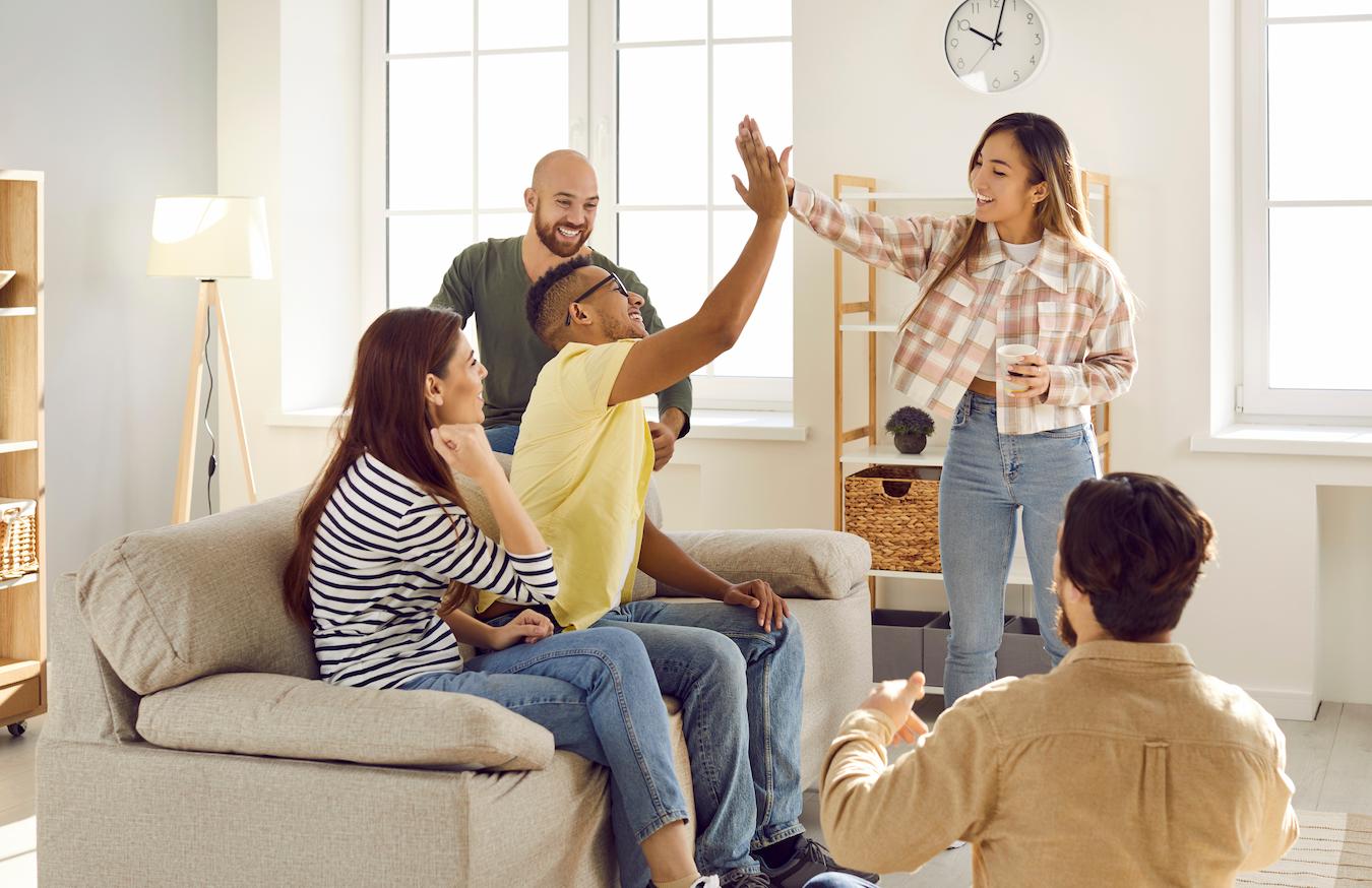 Five colleagues in bright room two sit on couch and two high five