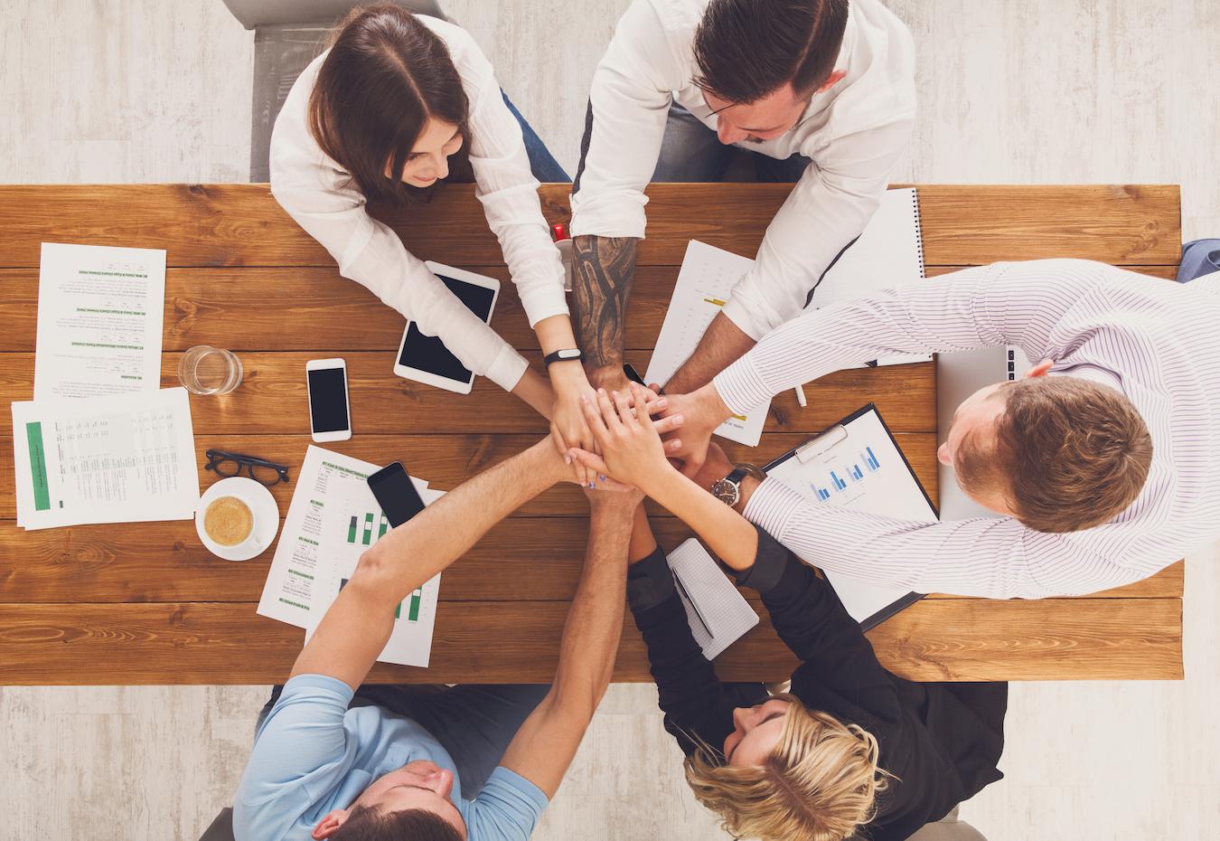 Five coworkers put their hands together over a long wooden table with phones and papers on it