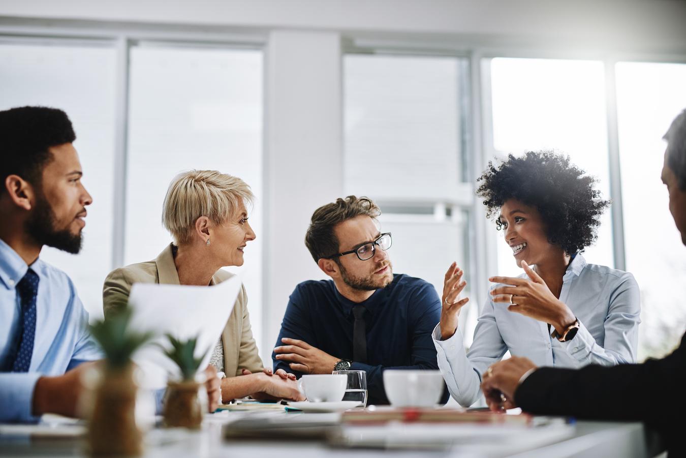 Five members of a corporate team sit at a desk in a bright office and create a plan to improve workplace culture for the staff at their business