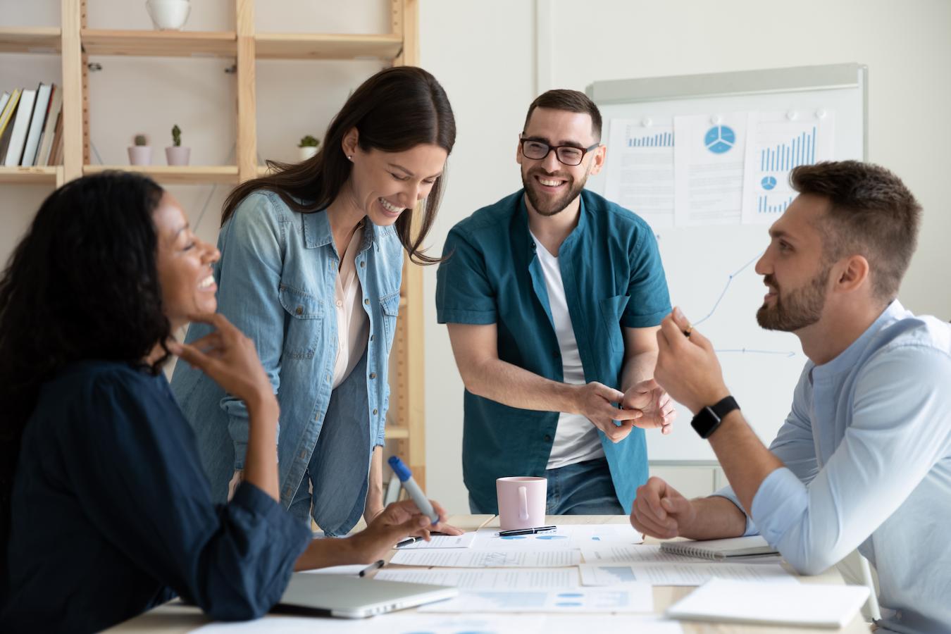 Four colleagues wearing varying shades of blue sit and stand around a table smiling and laughing as they review a very exciting upcoming marketing strategy