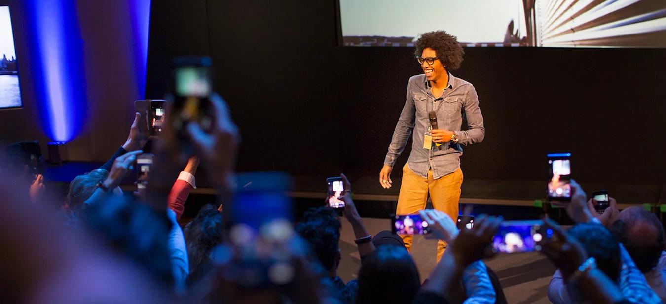 man speaking on stage while smiling with audience holding their phone audience confidence speech message audience speakers event audiences message audience