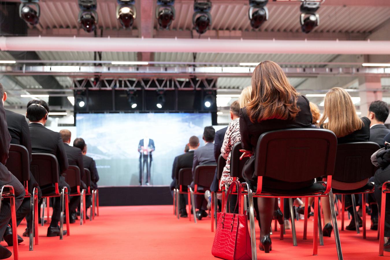 People sit in chairs facing a stage and listen to a speaker present in a room with red carpeted floors and a red purse in the foreground