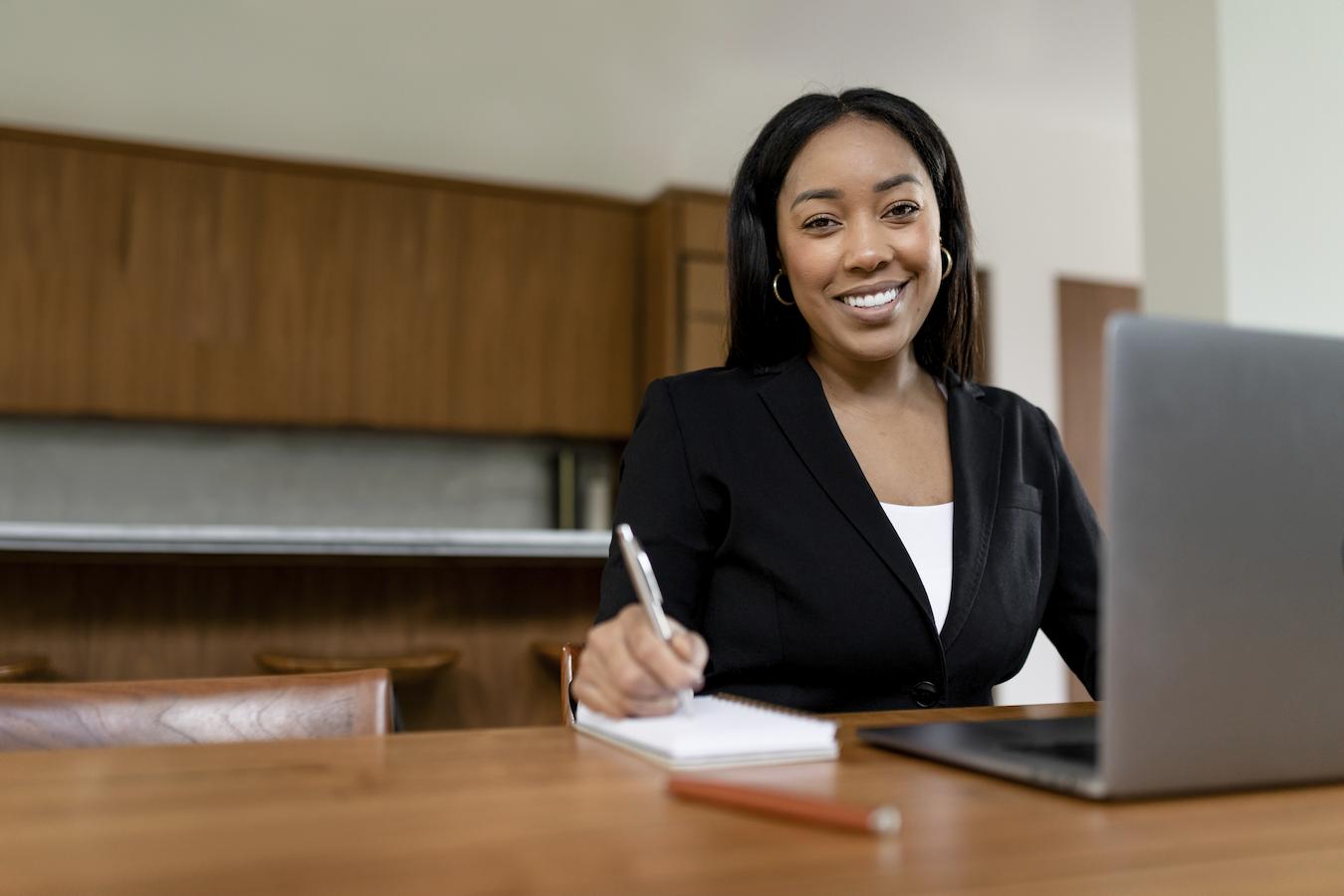 Person in black blazer sits at wooden desk in front of a laptop and puts pen to paper