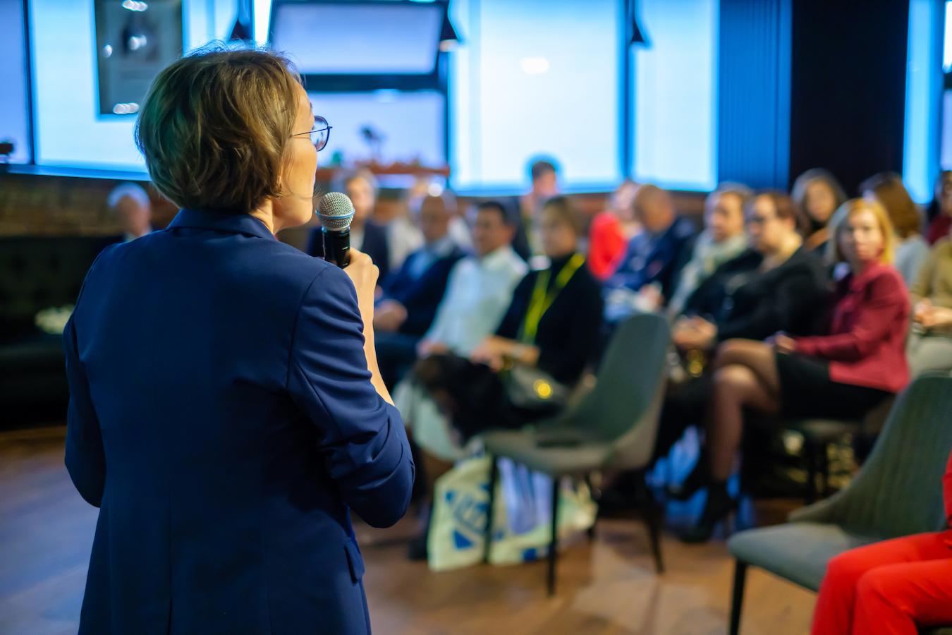 Person in blue business suit makes presentation and stands on small stage and faces audience excited to hear them talk while holding microphone