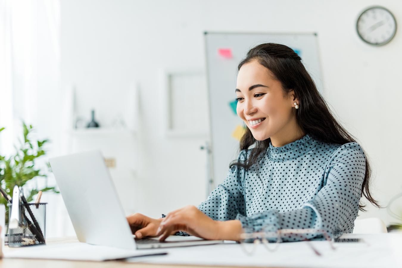 Person in blue dotted blouse smiles as type they on their computer in a bright office
