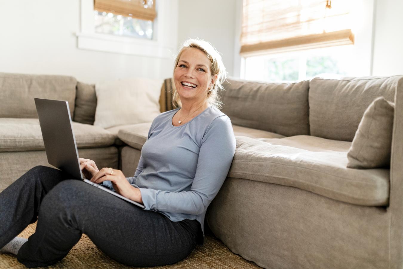 Person in blue top and gray pants smiles and sits on floor in front of couch with laptop