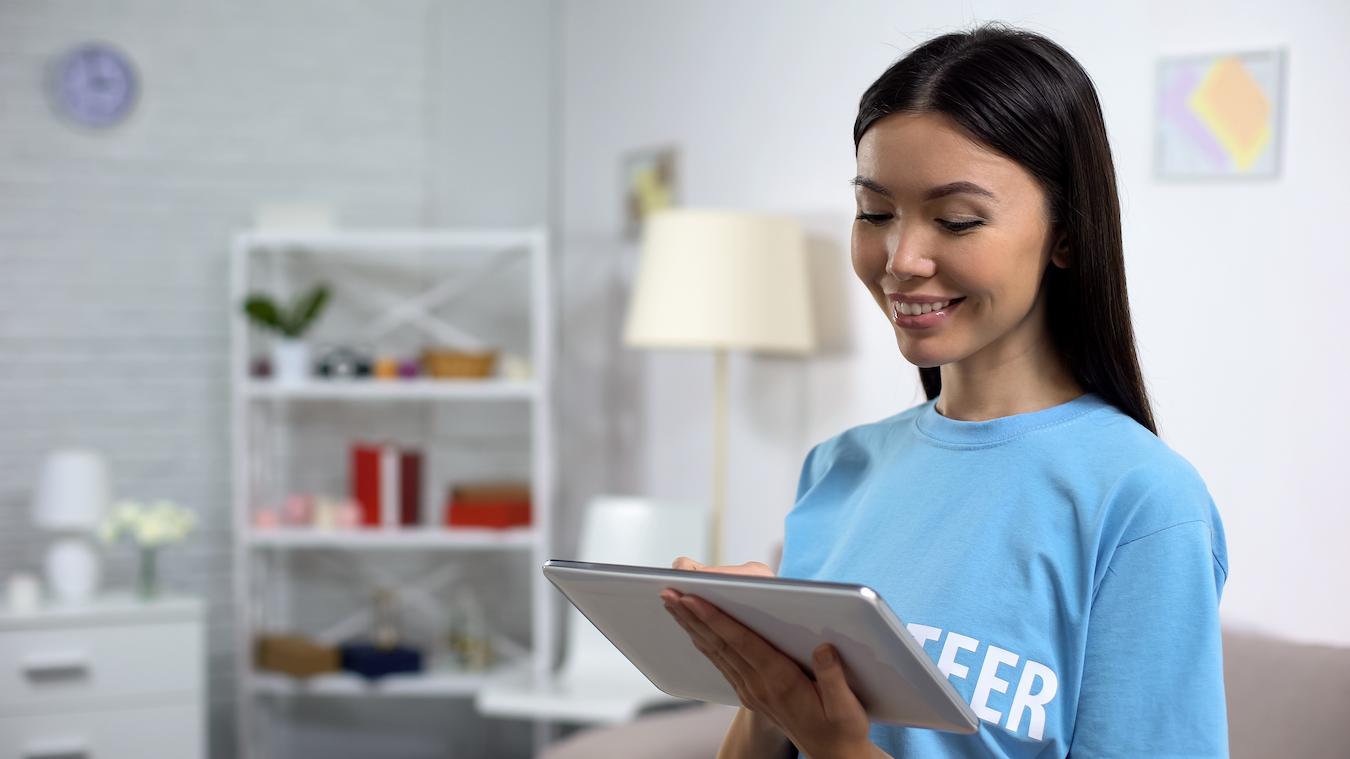 Person in blue volunteer t shirt smiles and works on their tablet in a bright office