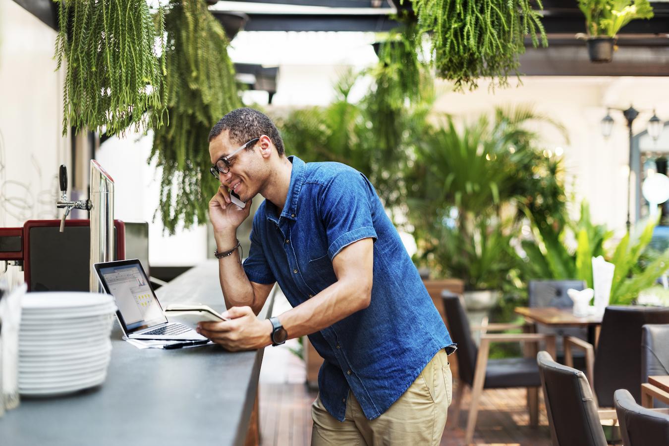 Person in denim button down and glasses talks on phone and works on laptop at a restaurant