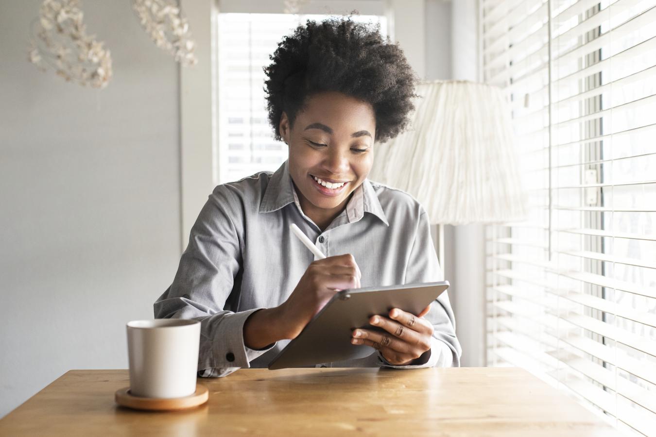 Person in gray button down sits at wooden table and works on tablet with stylus as coffee sits in front of them