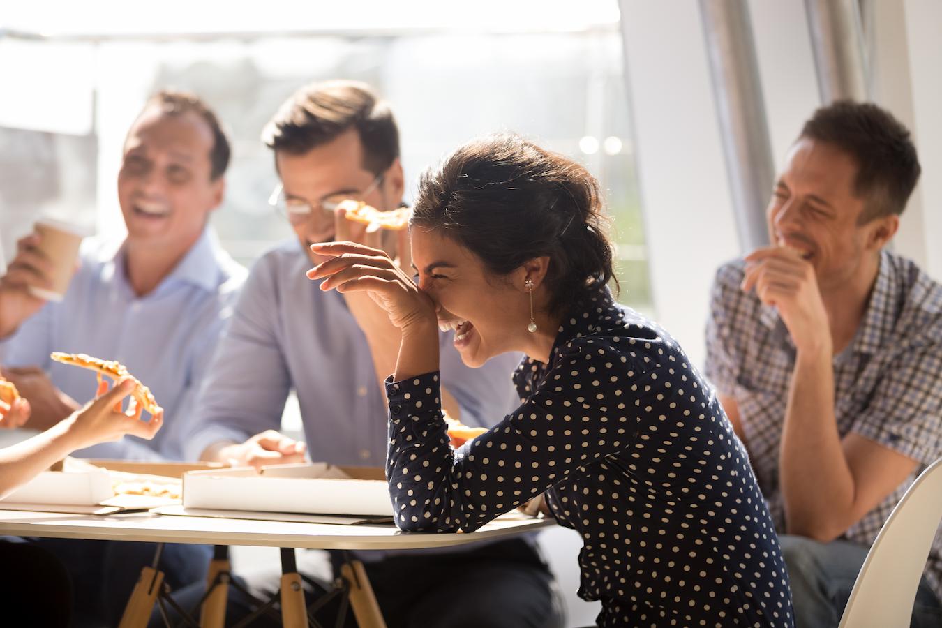 Person in polka dotted blouse laughs at a table with colleagues as they eat pizza and sit in a sunny office conference room