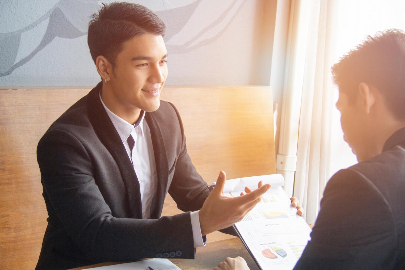 Person is reviewing a piece of paper with charts and graphs on it in a sunny office that has white curtains a wooden table and gray walls