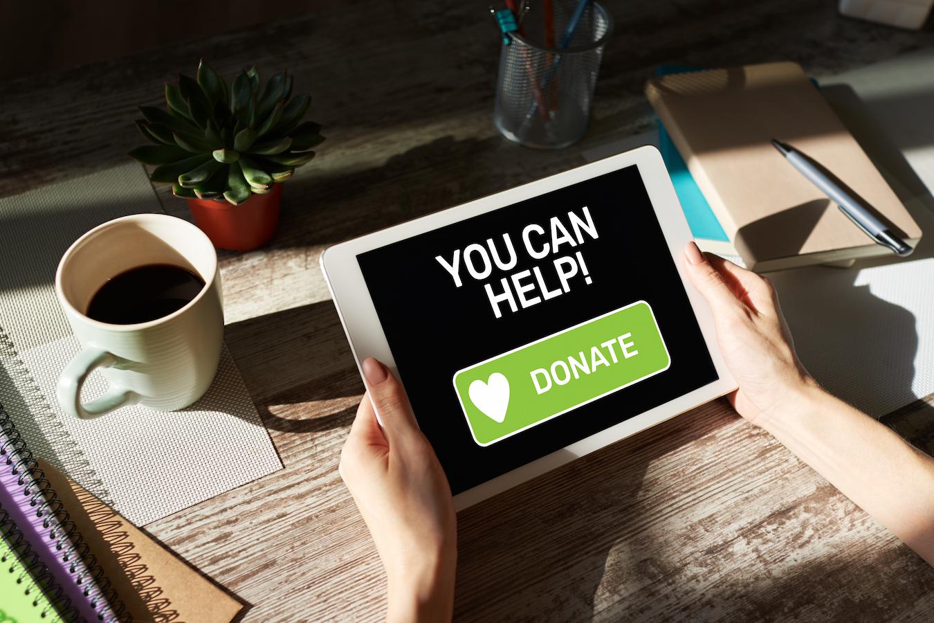 Person sits at wooden desk with a mug of coffee and a tablet they can use to donate to their favorite organization