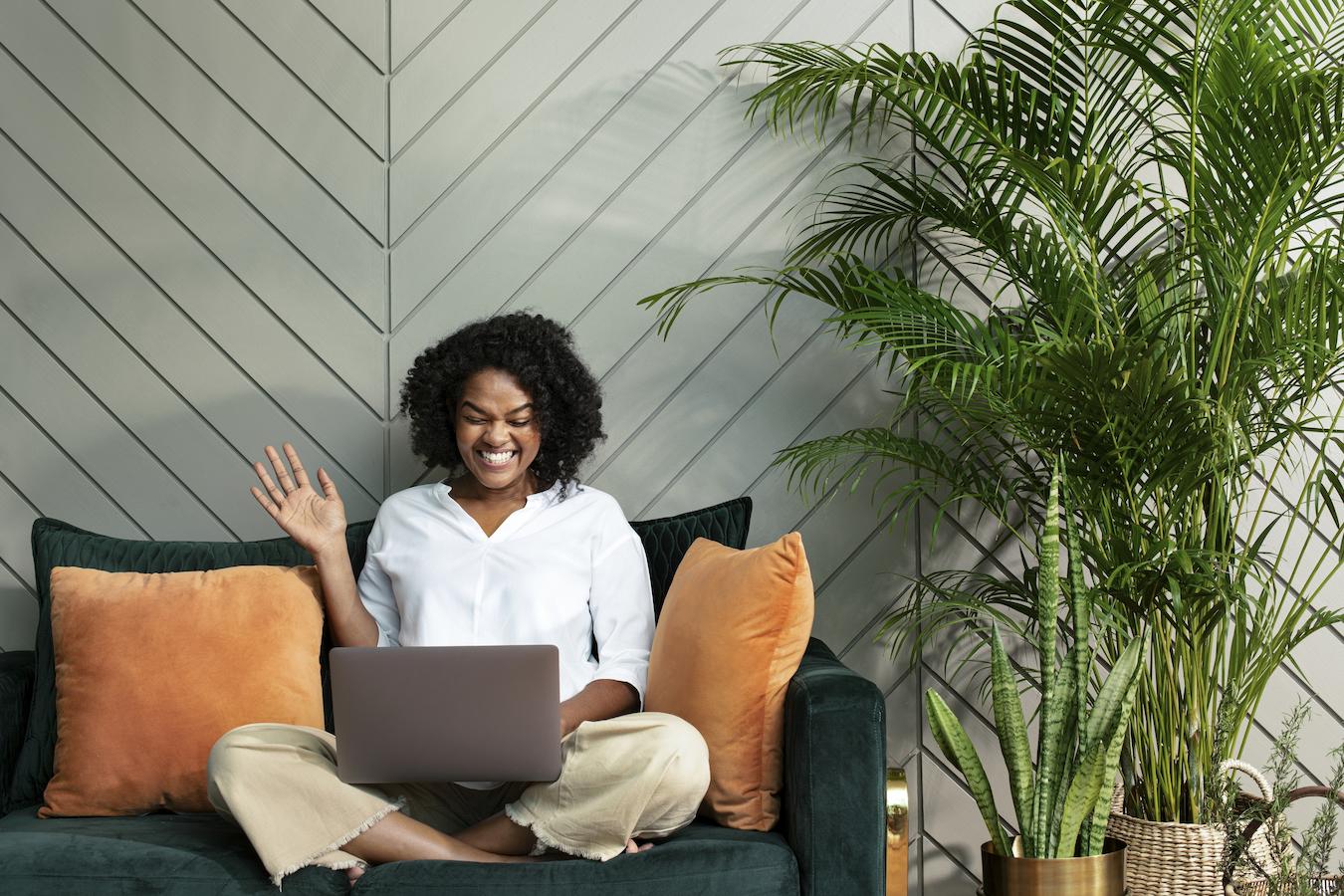 Person sits crossed legged on green couch and smiles at laptop screen