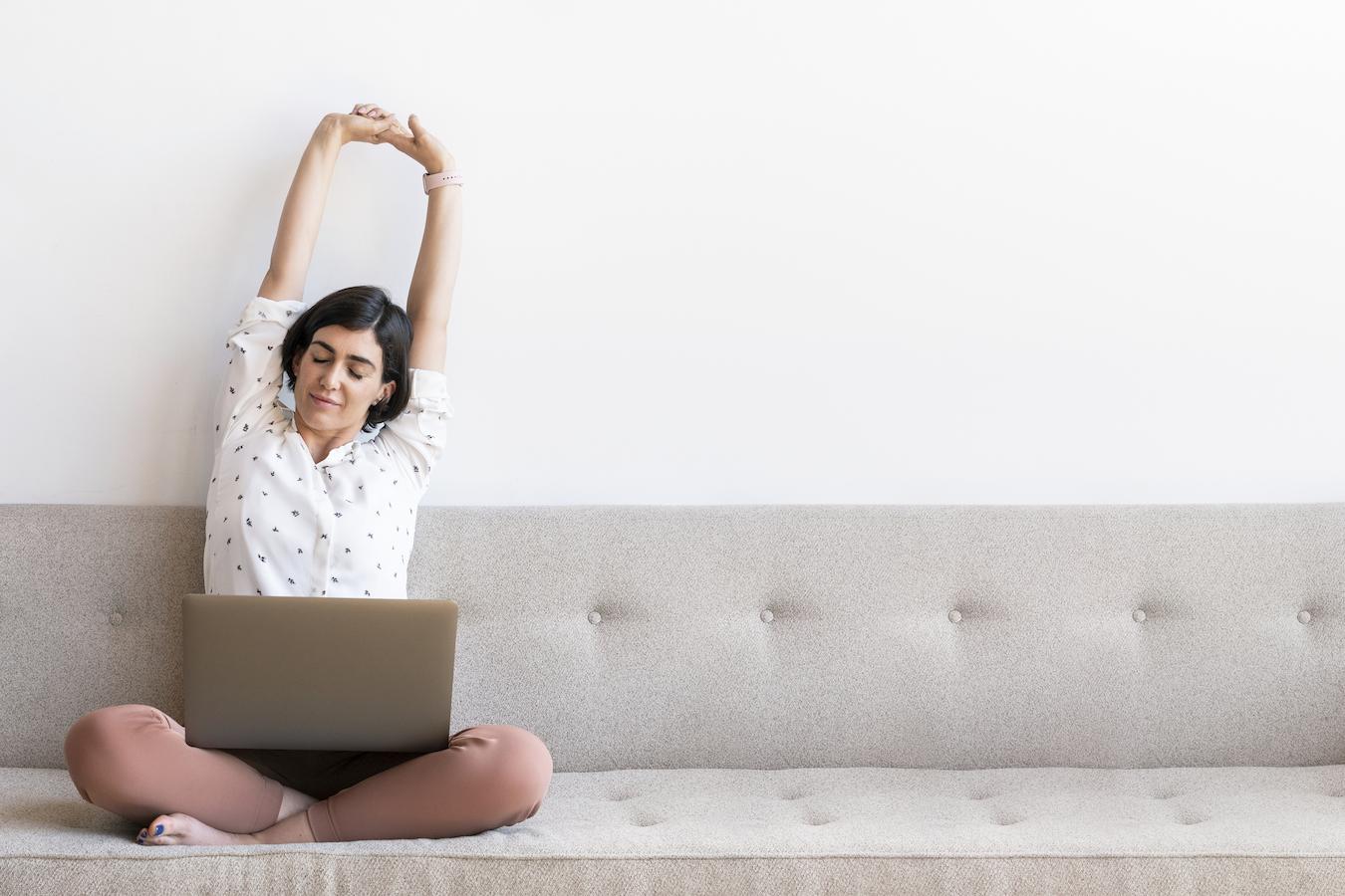 Person sits on gray couch and stretches while laptop sits in their lap
