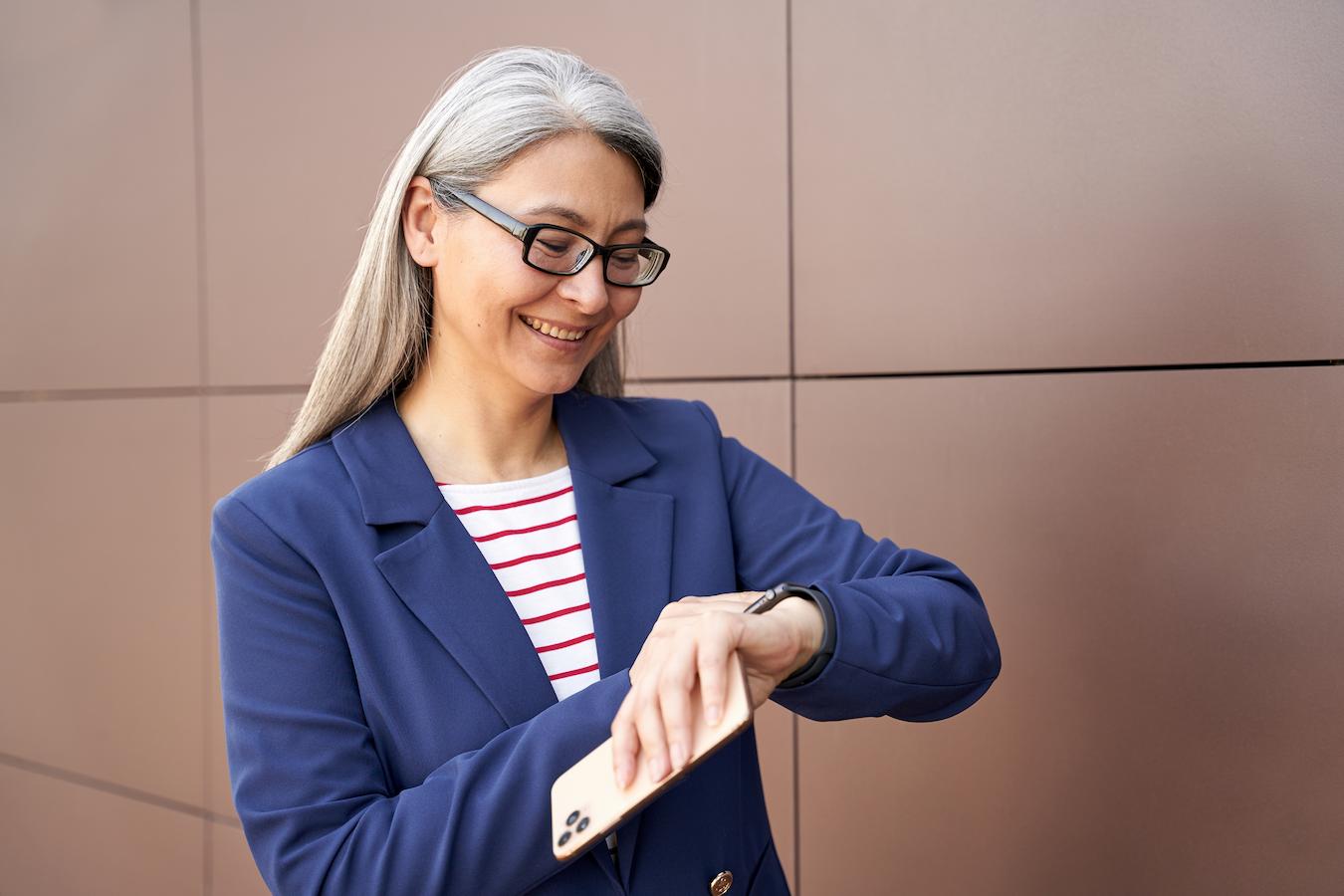 Person wearing blue blazer and white and red striped shirt smiles and looks at their watch while standing in front of a brown wall