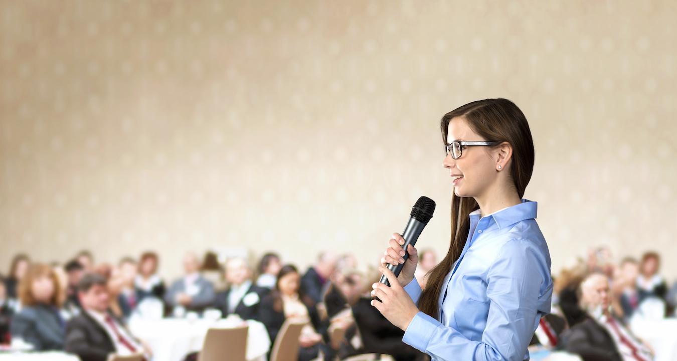 Person with brown hair in blue button down shirt makes speech to group of people excited to listen to their message
