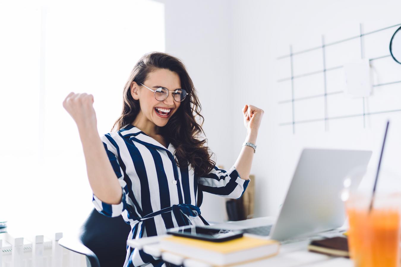 Person with long brown hair in blue and white striped top grins and raises their arms happily in front of a computer in a bright office
