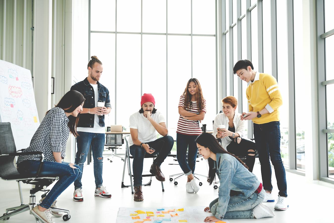 Seven coworkers in a white sunny office focus on completing a complex team building activity that involves colorful post it notes on the floor