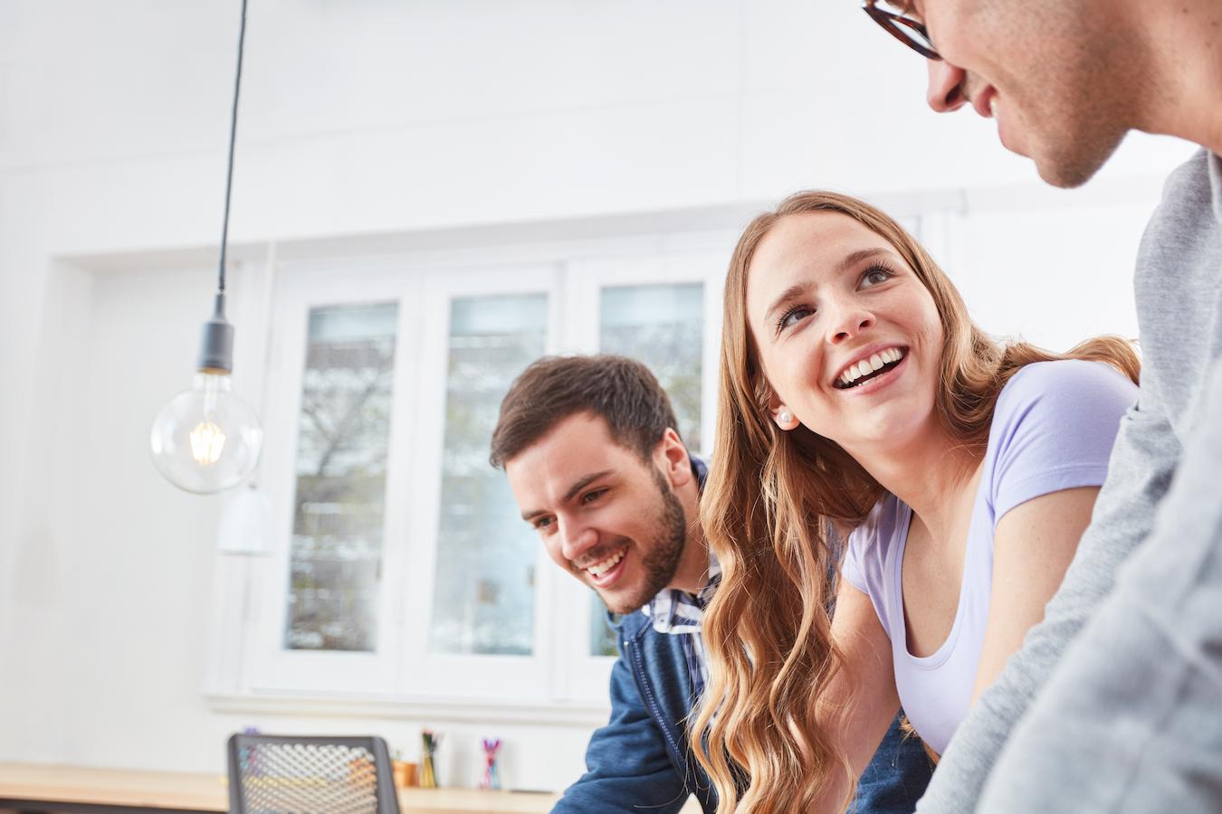 Three colleagues smile as they lean over a desk in a bright sunny office and look at something that is exciting