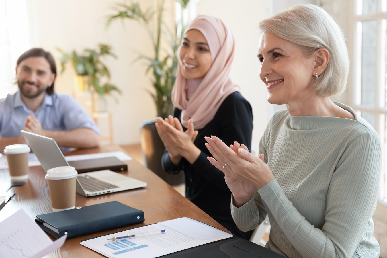 Three people clap and smile while sitting at conference table with papers on it