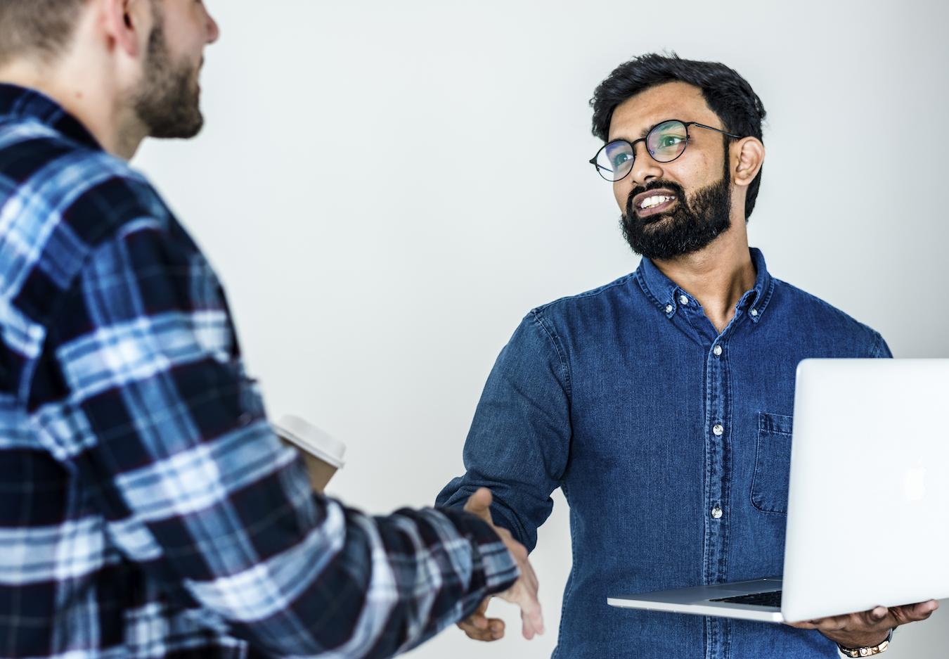 Two people in blue button downs shake hands as one holds a laptop